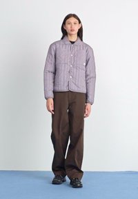 Vaatteena on harmaa pääntiellä varustettu tikkitakki, jossa on napitus ja kaksi sivutaskua, yhdistettynä ruskeisiin housuihin ja mustiin lenkkareihin.