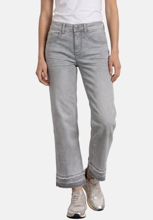 Kvinde iført lysegrå cropped jeans med frynsede kanter, hvide sneakers og en hvid ærmeløs top, stående mod en hvid baggrund.