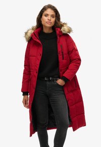 Abrigo largo rojo acolchado con capucha con borde de piel, bolsillos frontales con cremallera y diseño acolchado. Lleva puesto un suéter negro y jeans.