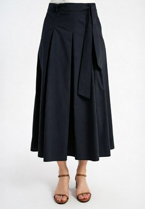 Granatowa midi spódnica z plisami i wiązaniem w talii z boku, noszona z brązowymi sandałami na obcasie z odkrytymi palcami i paseczkiem wokół kostki na białym tle.