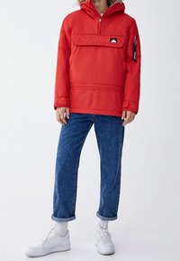 Persona che indossa una giacca a cappuccio rossa con tasca frontale, jeans blu arrotolati, calze bianche e sneaker bianche su uno sfondo bianco.