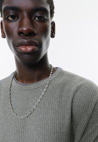Grå tekstureret sweater med rund hals, lagdelt over en blå skjorte. En sølvkæde er synlig omkring halsen.