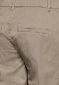Primo piano di pantaloni beige che mostrano la tasca posteriore con cuciture a welt e passanti per cintura sulla vita.