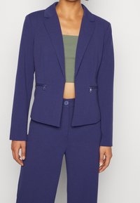 Fialový upravený blazer s ostrým límcem, který má dvě kapsy na zip. Materiál vypadá hladce a strukturovaně, v kombinaci s přiléhavým zeleným topem.