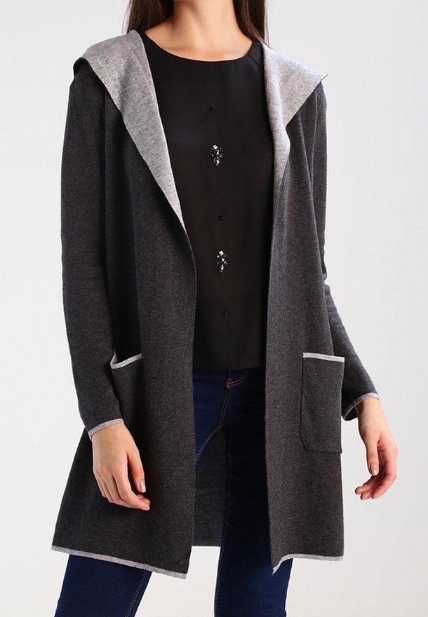 Femme portant un cardigan à capuche gris foncé ouvert devant avec un liseré gris clair sur un haut noir et un jean bleu foncé.