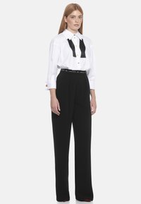 Camicia bianca con dettagli di cravatta nera a farfalla, bottoni dorati e maniche lunghe, abbinata a pantaloni neri a vita alta. Tessuto liscio.