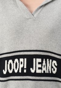Pull en tricot gris avec un col en V, comportant une bande noire au niveau de la poitrine avec le texte blanc "JOOP! JEANS" et une texture subtile.