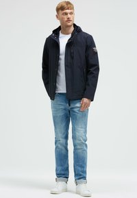 Granatowa kurtka z kapturem, z zamkiem z przodu, wieloma kieszeniami i regulowanymi mankietami. Nosi się ją na białej koszulce i zestawia z jasnoniebieskimi dżinsami.