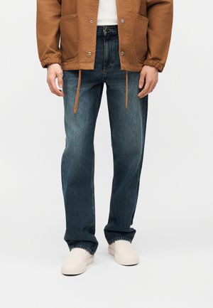 Oseba, oblečena v rjavo jakno, bele čevlje in široke temno modre kavbojke, stoji ob enobarvnem svetlem ozadju.