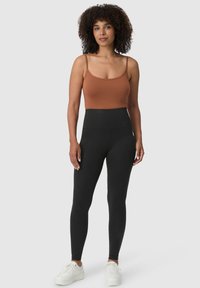 Bruine aansluitende tanktop met hoge taille zwarte legging. Model draagt witte sneakers. Glad textuur met minimalistisch ontwerp.