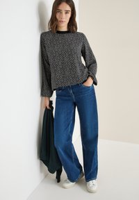 Top a maniche lunghe a righe in bianco e nero, vestibilità morbida, abbinato a jeans a gamba larga blu e sneakers bianche, mentre tiene un capo d'abbigliamento verde scuro.