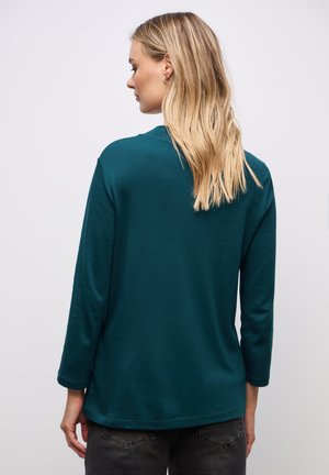 Maglia a maniche lunghe di colore teal con scollatura rotonda e vestibilità rilassata, caratterizzata da una texture morbida e un leggero drappeggio. L'orlo è leggermente curvato.