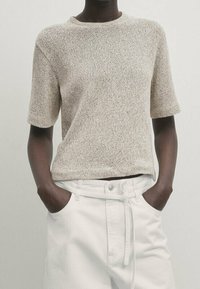 Beige lyhythihainen neulepaita pyöreällä kaula-aukolla, jossa on teksturoitu pinta. Päällä valkoiset korkeavyötäröiset housut, kädet taskuissa.