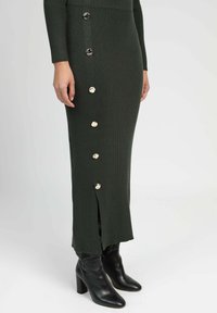 Robe midi en tricot verte avec une texture côtelée, boutons dorés sur le côté et fente à l'ourlet, associée à des bottes noires à talons.