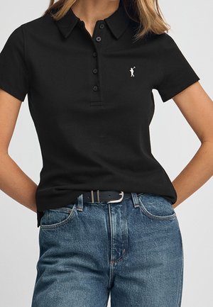 Kobieta ubrana w czarną polo z krótkim rękawem z małym białym logo golfisty, włożoną w niebieskie dżinsy z wysokim stanem i czarnym paskiem.