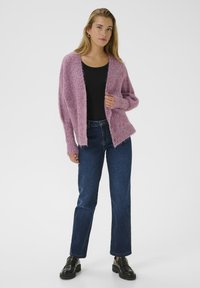 O femeie îmbrăcată cu un cardigan purpuriu pufoasă peste un top negru, blugi și pantofi cu talpă de piele negri, stând în fața unui fundal deschis simplu.