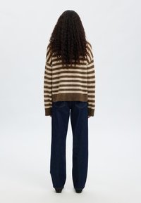 Barna és bézs csíkos kötött pulóver, laza szabással, bordázott mandzsettákkal, sötétkék bő szárú farmerrel párosítva. Az anyag puha és kényelmes hatású.