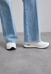 Weiße Sportschuhe mit glatter Textur, silbernen Akzenten an der Ferse und einer schwarzen Sohle, kombiniert mit hellblauen Schlagjeans.
