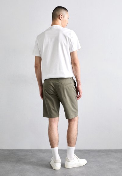 T-shirt blanc à manches courtes, shorts vert olive et baskets blanches. Les shorts sont de longueur genou avec des poches. Le modèle se tient de dos.