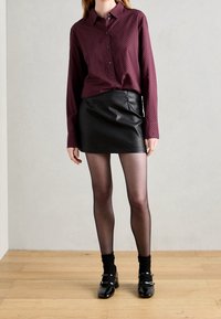 Camisa burdeos de botones con un patrón de puntos, combinada con una mini falda de cuero negra. El modelo lleva medias negras y zapatos negros brillantes.