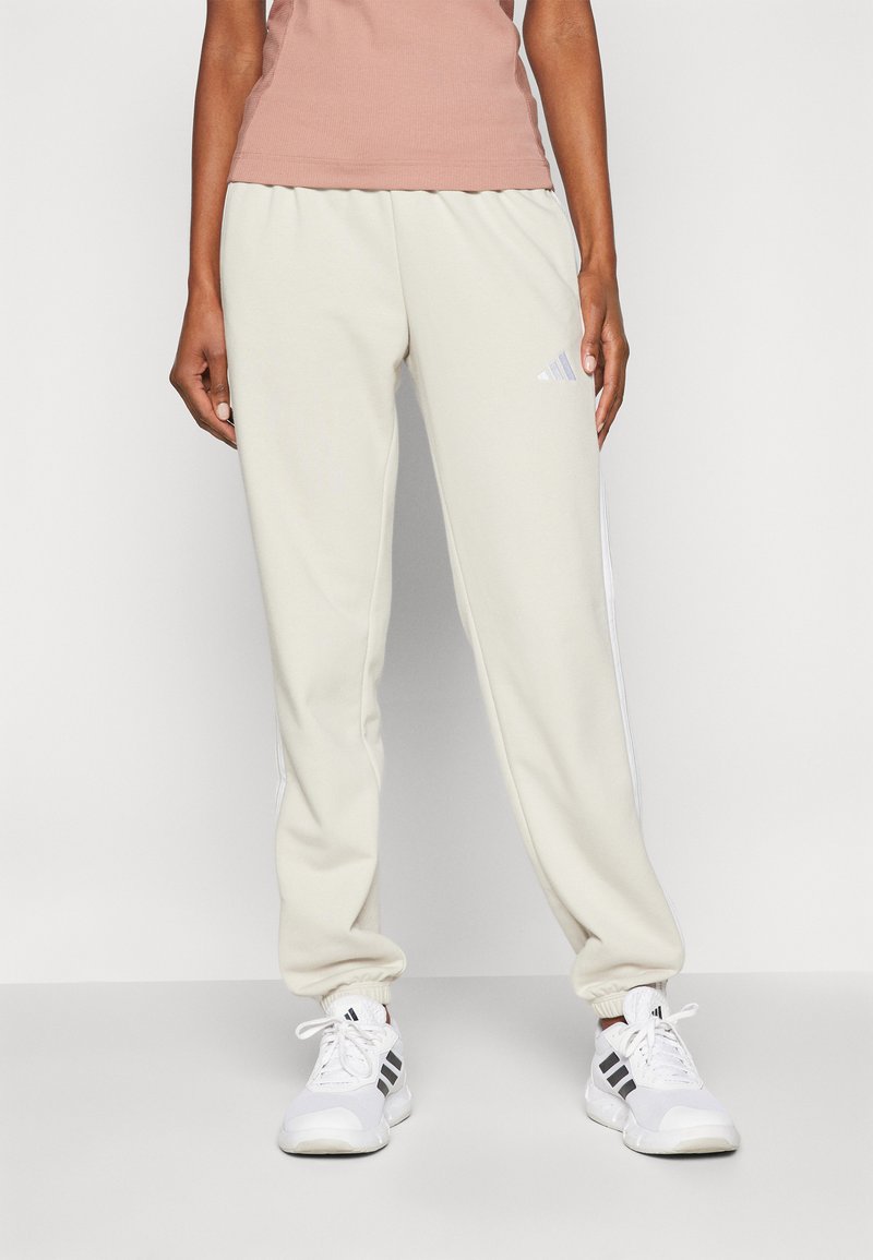 Lys beige joggebukser med elastiske mansjetter, med en hvit stripe nedover sidene og en subtil logo på låret. Laget av mykt stoff.