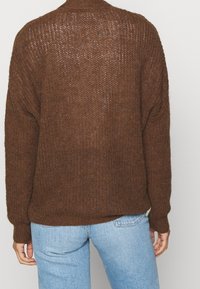 Volný hnědý pletený svetr s texturovaným vzorem, žebrovanými manžetami a vysokým límcem, nošený přes světle modré džíny.