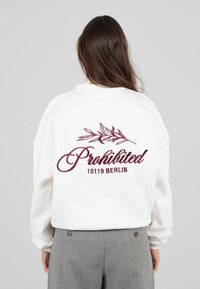 Donna con lunghi capelli castani che indossa una felpa bianca con testo in bordeaux "Proibito 10119 Berlino" e un design di foglia, abbinata a pantaloni grigi.
