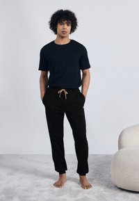 Crna majica kratkih rukava u kombinaciji s crnim trenirkama koje imaju vezanje i svijetlosmeđu uže. Goli noge na teksturiranoj površini.