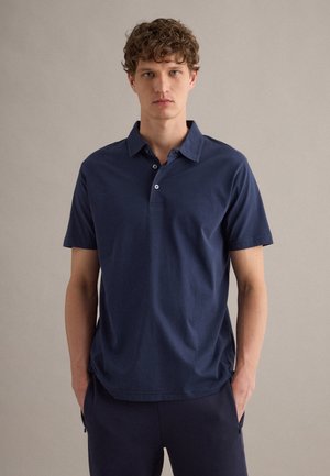 Jeune homme aux cheveux bouclés portant un polo bleu foncé à manches courtes et un pantalon foncé, debout devant un fond uni clair.