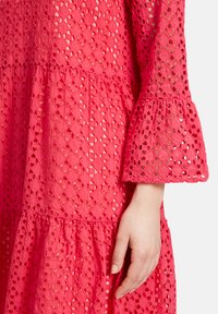 Rosa Spitzenkleid mit einem Diamantmuster, ausgestattet mit Glockenärmel und einem gestuften Design. Der Stoff ist leicht und hat eine strukturierte Oberfläche.