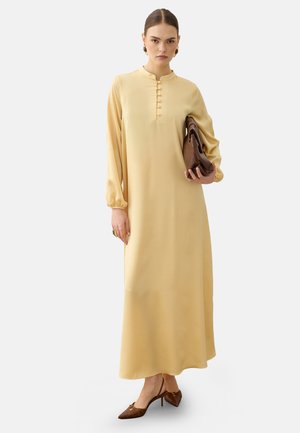 Frau trägt ein langes beiges Kleid mit Knopfdetails, hält eine braune Clutch und trägt braune spitze High Heels, steht vor weißem Hintergrund.