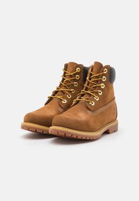Hnedé semišové ankle boots so žltými šnúrkami, zlatými očkami a čiernymi akcentmi na podpätkoch. Pevná gumová podrážka s textúrovaným dezénom pre lepšiu trakciu.