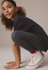 Sudadera azul marino, leggings grises y zapatillas blancas de caña alta con detalles en negro. Cabello en rizos sueltos, pose agachada contra un fondo neutro.