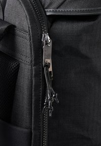 Črn tkaninski nahrbtnik z gladkim zadrgo in zaobljenimi zadrgnimi potegi, s stranskim žepom iz mrežastega materiala in minimalistično teksturo. Detajli v temnem kovinskem odtenku.