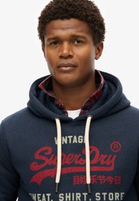 Marineblauwe hoodie met crèmekleurige koordjes en rode logotekst. Gelaagd met een geruit flanellen shirt eronder, met een witte kraag.