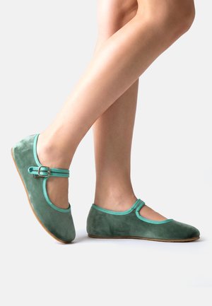 Beine, die grüne Wildleder-Mary-Jane-Ballerinas mit hellgrüner Einfassung und Knöchelriemen tragen, auf weißem Hintergrund posiert.