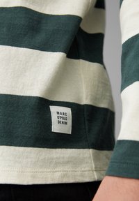Nahaufnahme eines gestreiften grünen und weißen Gewebes mit einem eingestickten MARC O'POLO DENIM Etikett in der Nähe des Saums, getragen etwas oberhalb einer schwarzen Hose.