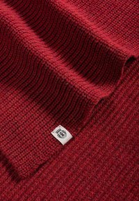 Red textured knit fabric with a ribbed pattern, featuring a small label in the bottom corner with black and white details.
