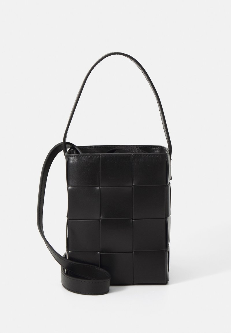 Petit sac bandoulière en cuir noir avec motif carré tressé et longue sangle sur fond uni clair.