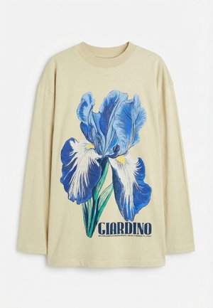 Chemise beige à manches longues avec un grand motif fleur d'iris bleu et blanc et le texte "GIARDINO" avec le nom de l'artiste et le lieu en dessous.