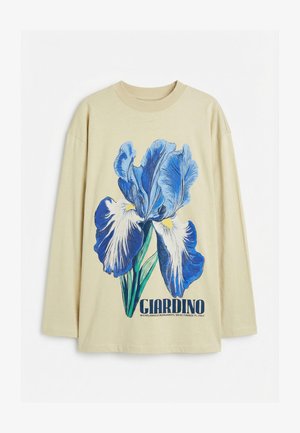 Chemise beige à manches longues avec un grand motif fleur d'iris bleu et blanc et le texte "GIARDINO" avec le nom de l'artiste et le lieu en dessous.
