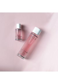 Två transparenta flaskor med rosa blommig vatten, en större och en mindre, båda med vita lock och svart text på framsidan, på en ljusrosa bakgrund.