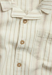 Knoopshirt met verticale strepen in beige en crème. Heeft een zachte, gestructureerde stof en houten knopen met een zak aan de linkerkant.