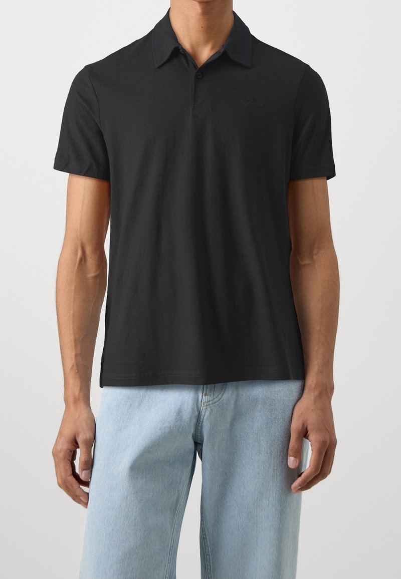 Homme portant un polo noir uni à manches courtes et un jean bleu clair, debout devant un fond uni.