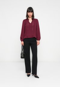 Blouse bourgogne à manches longues, à texture plissée et décolleté en V ; associée à un pantalon noir et des talons. Le mannequin tient un petit sac noir.