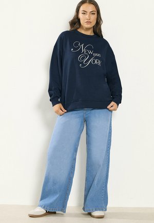 Frau trägt einen marineblauen Pullover mit dem Aufdruck "New York 1998" und hellblaue Jeans mit weitem Bein, steht vor einem einfarbigen hellen Hintergrund.
