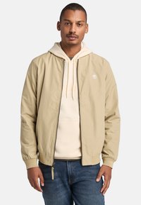 Chaqueta beige con una textura suave, puños acanalados y cremallera frontal. Combinada con una sudadera color crema y unos vaqueros. Logotipo en la chaqueta.
