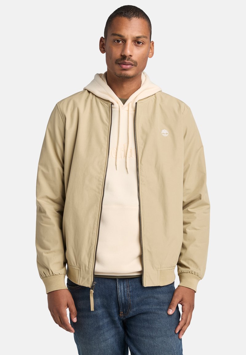 Chaqueta beige con una textura suave, puños acanalados y cremallera frontal. Combinada con una sudadera color crema y unos vaqueros. Logotipo en la chaqueta.