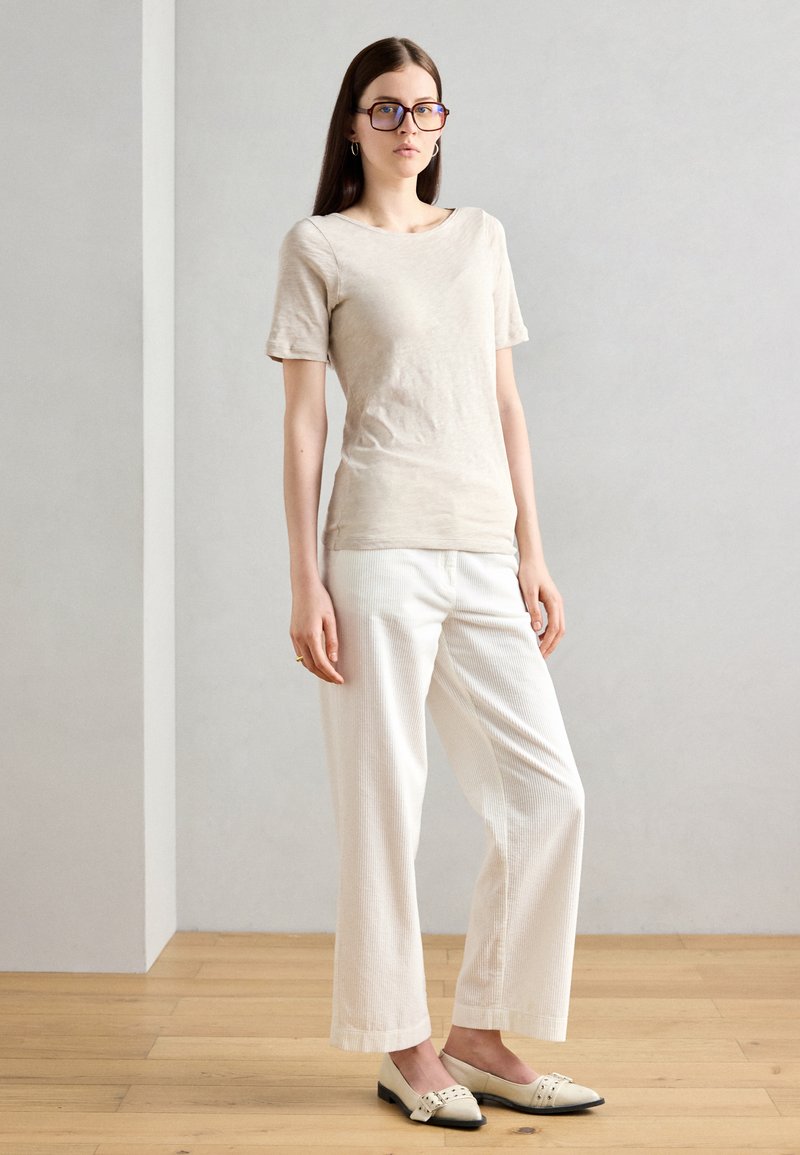 Beige Kurzarmhemd kombiniert mit weißen, strukturierten weit geschnittenen Hosen und flachen Schuhen mit einer schwarzen Zehenkappe und dekorativen Akzenten.