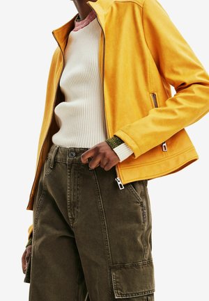 Person wearing a yellow zip-up jacket over a white ribbed sweater and dark green cargo pants with side pockets.